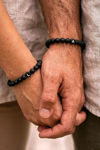 Pärchen-Armbänder mit Wunschbuchstaben (L und A) aus Naturstein-Onyxperlen, getragen am Handgelenk eines Paares in Leinenkleidung.